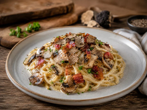 Wide view of creamy spaghetti with mushrooms, bacon and shaved black truffle, served on a ceramic plate in a rustic kitchen setting.