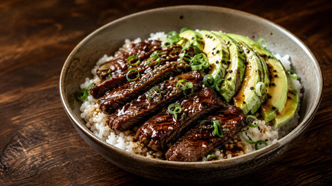 Wide view of a teriyaki-inspired beef rice bowl with avocado, glazed ribeye strips, Japanese rice, sesame, green onions and black truffle oil.